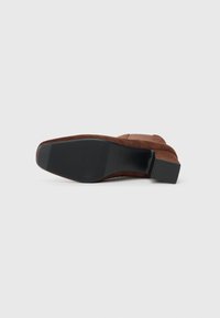 Chaussure à talon en daim marron avec un bout carré et une surface texturée. La semelle est en caoutchouc avec des rainures horizontales pour une meilleure adhérence.