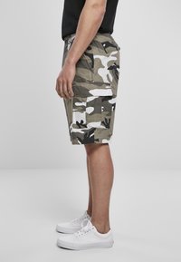Shorts cargo camouflage en vert olive, blanc et noir, avec plusieurs poches et un tissu texturé. Portés avec des baskets blanches.