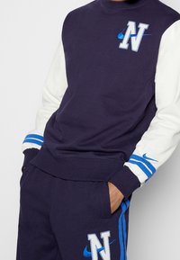 Marinblå sweatshirt med vita ärmar, prydd med broderat "N" och Nike-logga i blått. Ribbad mudd med blåa ränder, tillverkad av mjukt bomullsmaterial.