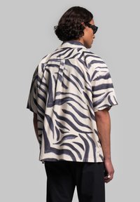 Camicia a maniche corte color crema con righe zebrate nere, caratterizzata da una vestibilità comoda, yoke posteriore e orlo dritto. Materiale in cotone morbido.
