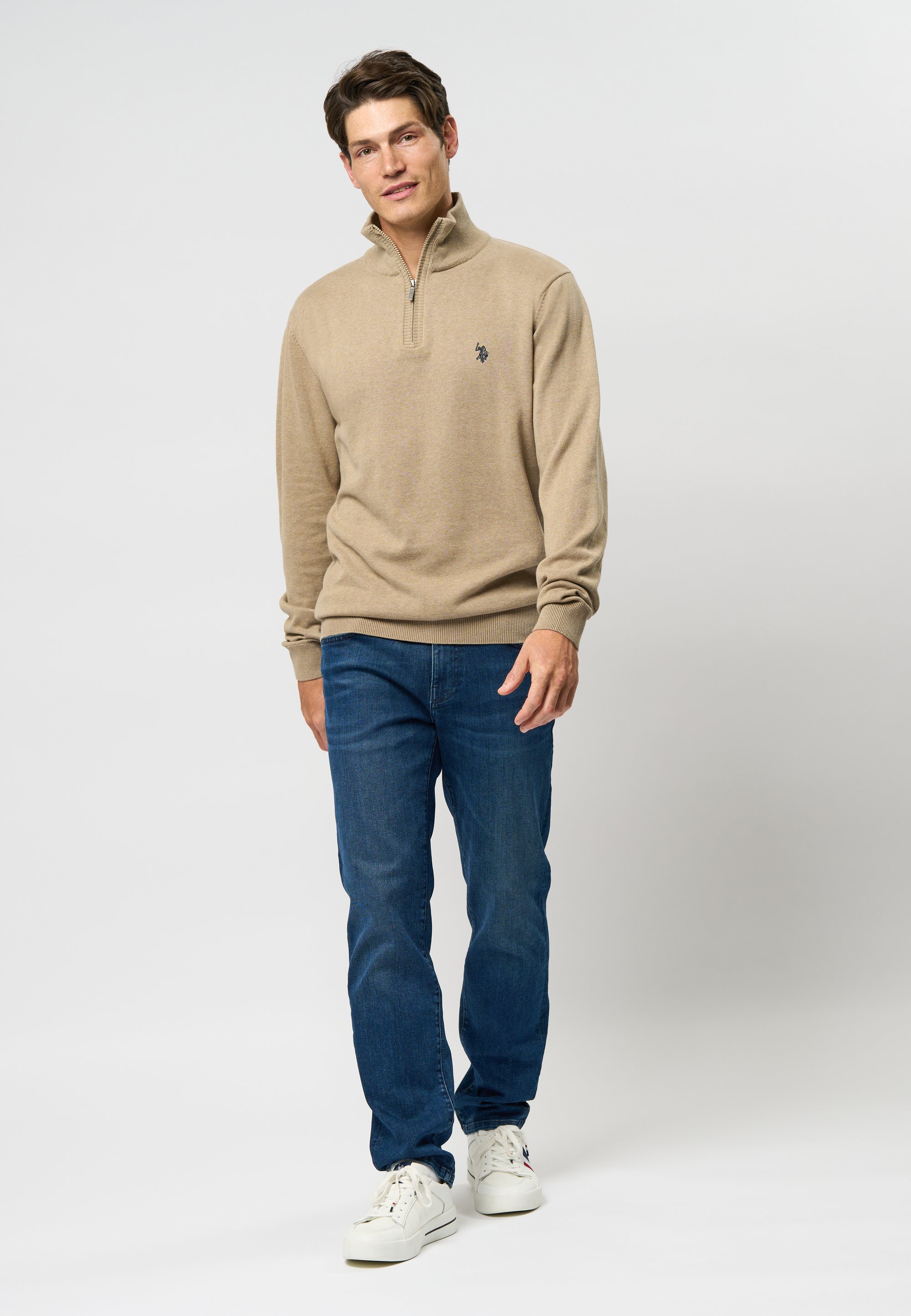 Man wearing a beige quarter-zip sweater, blue jeans, and white sneakers standing against a plain light background.