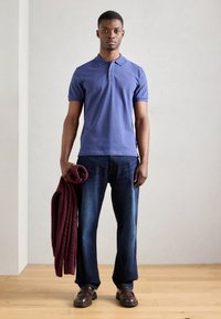 Polo shirt in cotone blu, jeans in denim scuro, scarpe in pelle marrone e un oggetto lavorato a maglia bordeaux tenuto nella mano sinistra. Design semplice.