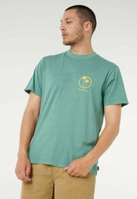 Uomo con maglietta verde con logo di palma gialla e pantaloni beige, che guarda a sinistra su uno sfondo neutro.