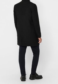 Zwarte wolmixjas met een hoge kraag, enkele rugventilatie en knopenmanchetten, gecombineerd met donkere broeken en zwarte schoenen.