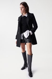 Manteau en laine noir à double boutonnage, pull noir, chemise blanche, short en cuir noir et bottes à talons noires montantes.