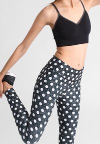 Sujeción deportiva negra con tirantes ajustables, combinada con leggings de cintura alta que tienen un fondo oscuro y grandes lunares blancos.