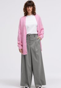 Pink knitted cardigan with ribbed edges, worn over a white t-shirt featuring embossed text. Gray pinstriped wide-leg trousers and sneakers.