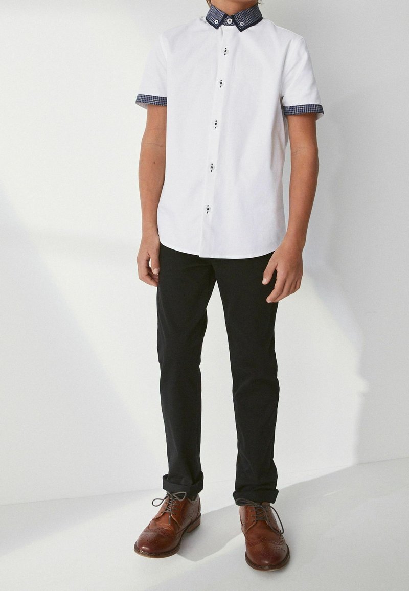 Garçon portant une chemise blanche à manches courtes avec col et poignets à motifs, pantalon noir et chaussures de ville marron, debout contre un mur blanc.
