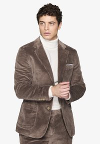 Brun kordfløjlsblazer med slag og to forlommer samt tre knapper. Båret over en hvid polo, glat tekstur.