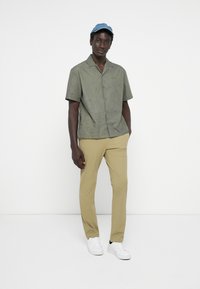 Camisa de manga corta verde oliva con patrón topográfico, combinada con pantalones beiges y zapatillas blancas, complementada con una gorra de denim clara.