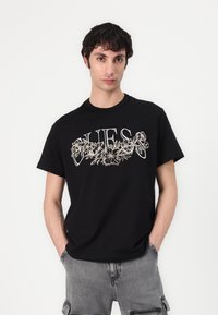 Black cotton t-shirt featuring a floral graphic design in white and beige. Crew neck, short sleeves, relaxed fit. Worn with gray shorts.