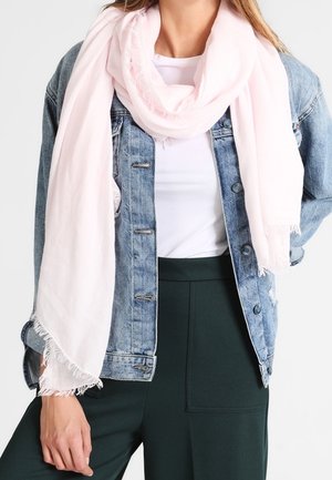 Femme portant une écharpe à franges rose clair sur une veste en denim bleue, un haut blanc et un pantalon foncé taille haute, la main reposant près de la hanche.
