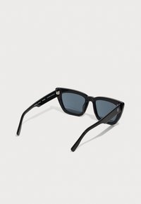 Ochelari de soare negri cu o formă triangulară, lentile întunecate și rame subțiri din plastic. Brațele sunt elegante, cu un detaliu subtil al logo-ului.