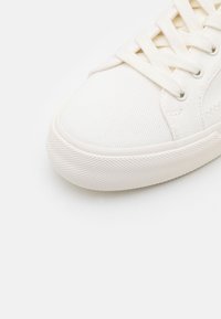 Białe trampki z płótna o teksturowanej powierzchni, okrągłym nosku, płaskimi sznurówkami i gumową podeszwą z diamentowym wzorem dla lepszej przyczepności.