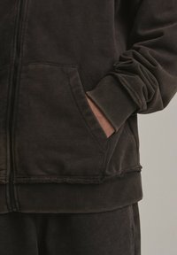 Ciemnobrązowa bluza z kapturem, zapinana na zamek, z ściągaczami na rękawach i kieszeniami. Charakteryzuje się miękką fakturą i postrzępionym dołem.