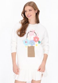 Fehér pulóver kosármintával, amely texturált rózsaszín, kék, sárga és fehér virágokat ábrázol, rózsaszín masni részlettel díszítve.