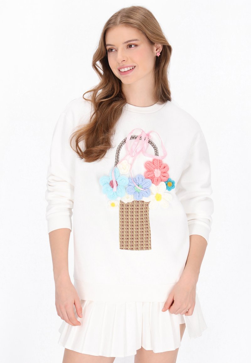 Fehér pulóver kosármintával, amely texturált rózsaszín, kék, sárga és fehér virágokat ábrázol, rózsaszín masni részlettel díszítve.