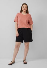 Oversized różowy t-shirt w odcieniu kurzu, czarne dopasowane szorty z kieszeniami oraz czarne sandały na paski. Prosty, luźny strój na modelce.