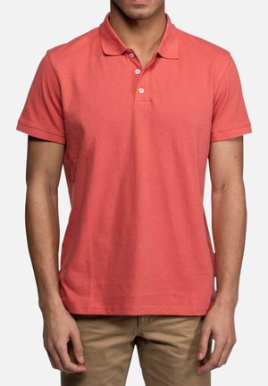 Homme portant un polo corail à manches courtes avec deux boutons, associé à un pantalon marron clair, debout sur un fond blanc.