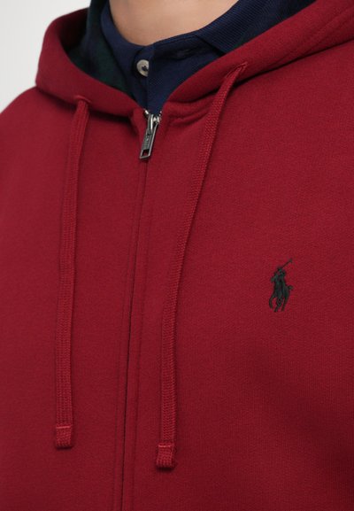 Hoodie vermelho com fecho éclair, feito de tecido macio, apresentando um capuz com cordão e um pequeno logotipo preto no peito.