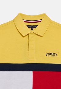 Κίτρινο πόλο πουκάμισο με γιακά, με οριζόντιες ρίγες σε ναυτικό μπλε και κόκκινο στο κάτω μέρος. Κεντημένο λογότυπο στην αριστερή πλευρά.