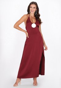 Burgundowa maxi sukienka na halterze z głębokim dekoltem w kształcie litery V i bocznym rozcięciem. Posiada biały kwiatowy akcent na piersi. Gładka satynowa faktura.