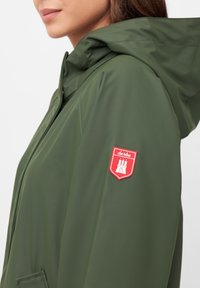 Giacca impermeabile verde con cappuccio, tasca laterale e un emblema rosso con il design di un castello sulla manica. Texture liscia e lucida.