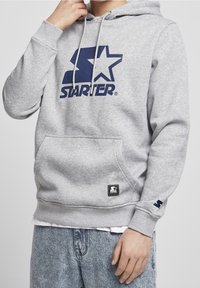 Persoon in grijze Starter hoodie met blauw logo en lichtblauwe spijkerbroek, hand geheven nabij het gezicht, vooraanzicht gecropt boven de mond.
