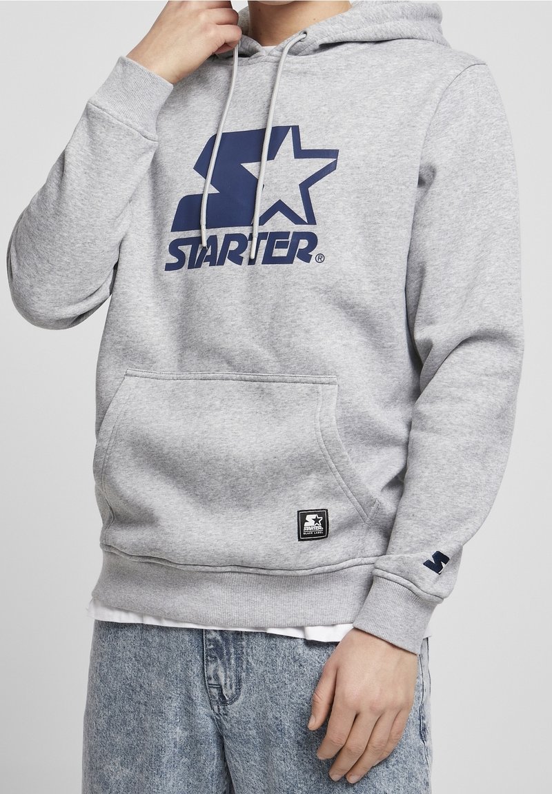Persoon in grijze Starter hoodie met blauw logo en lichtblauwe spijkerbroek, hand geheven nabij het gezicht, vooraanzicht gecropt boven de mond.