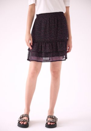 Vrouw draagt een zwarte en paarse rok met luipaardprint en rucheslagen, een wit topje en zwarte sandalen met zilveren gespen.