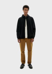 Zwarte zip-up hoodie met een trekkoord, gedragen over een wit shirt. In combinatie met een paar bruine broeken en zwarte schoenen, met een relaxte pasvorm.