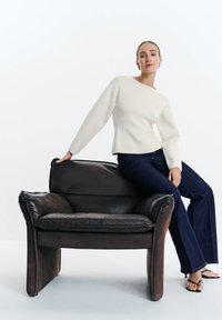 Femme en pull blanc et jean foncé assise sur l'accoudoir d'un fauteuil en cuir marron foncé sur fond blanc.