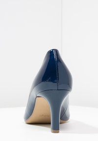 Marinblå lackläder högklackad sko med spetsig tå. Har en smal klack och en slät, glänsande yta. Modellnumret synligt på sulan.