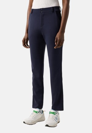 Personne portant un pantalon droit marine, des baskets blanches avec des lacets verts, et un haut blanc sans manches, debout devant un fond uni.