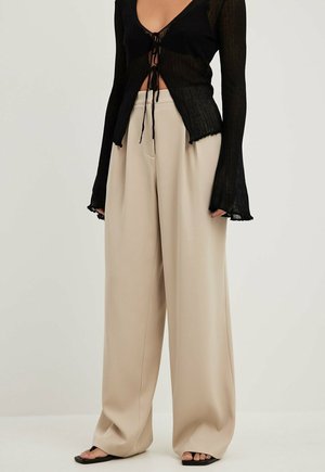 Sort langærmet crop top med snører foran, kombineret med højtaljede beige bukser med vide ben og sorte sandaler med åbne tæer.