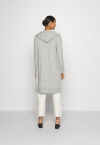 Langer grauer Cardigan mit Kapuze, taillierten Ärmeln und seitlichen Schlitzen, kombiniert mit weißen, kurzen Hosen und schwarzen, hohen Sandalen.