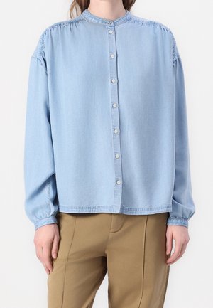 Femme portant une chemise bleu clair à manches longues boutonnée avec un col montant et un pantalon beige, debout devant un fond blanc uni.