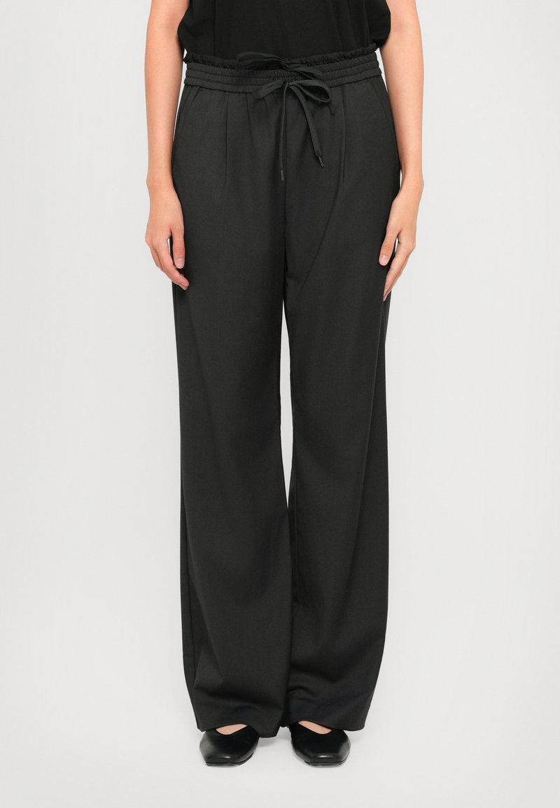 Personne portant un pantalon noir ample taille haute avec un cordon à la taille et des chaussures plates noires, debout devant un fond uni.