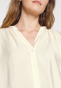Blusa amarula clara com botão, featuring um decote em V, ombros franzidos e textura rugosa. Botões feitos de plástico transparente.