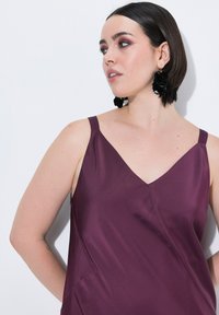 Burgundowa satynowa koszulka na ramiączkach z dekoltem w kształcie litery V i symetrycznymi ramiączkami, z gładką fakturą i ukośnymi szwami. Stylizowana z czarnymi kolczykami.