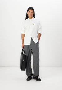 White button-up shirt with a chest pocket and yellow logo, paired with grey pinstripe trousers, holding a black tote bag, wearing black shoes.