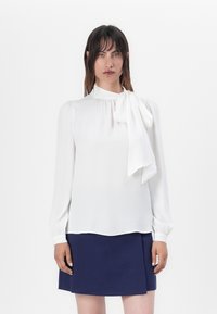 Valkoinen pitkähiha blouse korkealla kauluksella ja suurella rusettidetaljilla, yhdistetty tummansiniin hameeseen. Sujuva kangas ja rento muotoilu.