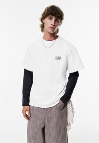 Hvid bomuld t-shirt lagdelt over en marineblå langærmet skjorte med et lille logo-patch. Grå stribede wide-leg bukser. Sølv halskæde og ring.