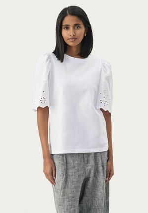 Weiße Baumwollbluse mit kurzen, gepufften Ärmeln, die mit detaillierter Lochstickerei und gewellten Kanten verziert ist. Kombiniert mit grau strukturierten Hosen.