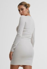 Langermet, tettsittende kjole i skinnende lys grått stoff. Har rund hals og design som fremhever silhuetten.