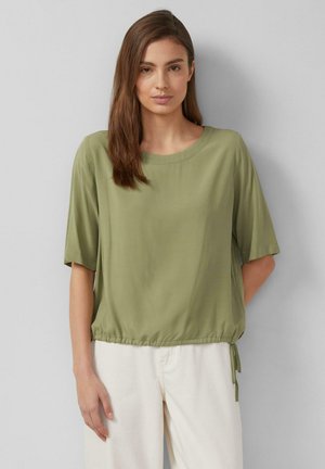 Kobieta z prostymi brązowymi włosami, ubrana w oliwkowozieloną bluzkę z krótkim rękawem z regulowanym dołem i kremowe spodnie, stojąca na szarym tle.