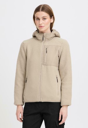 Kobieta w beżowej polarowej kurtce zapinanej na zamek z kapturem i kieszeniami na zamek, stojąca na białym tle.