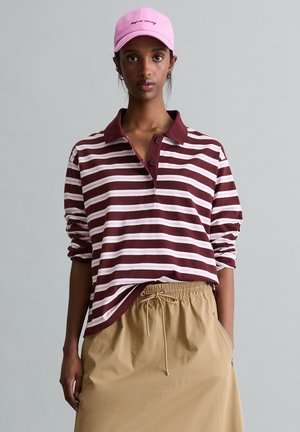 Vrouw met een roze pet, een bordeauxrood-wit gestreept poloshirt met lange mouwen en een beige rok met trekkoord tegen een effen grijze achtergrond.