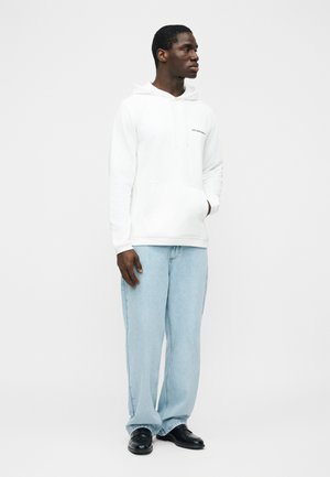 Moški, ki nosi belo kapuco z vrečko spredaj, svetlo modre široke kavbojke in črne čevlje, stoji pred preprostim belim ozadjem.