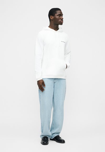 Homme portant un sweat à capuche blanc avec une poche avant, un jean bleu clair à jambes larges et des chaussures noires, debout devant un fond blanc uni.